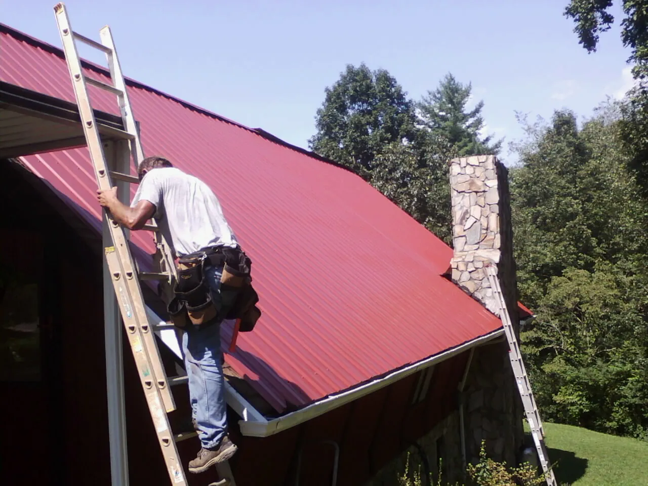 Roofer on a ladder beside a red metal roof and stone chimney — Soffit & Fascia Repair by Roofing Brothers in Wever